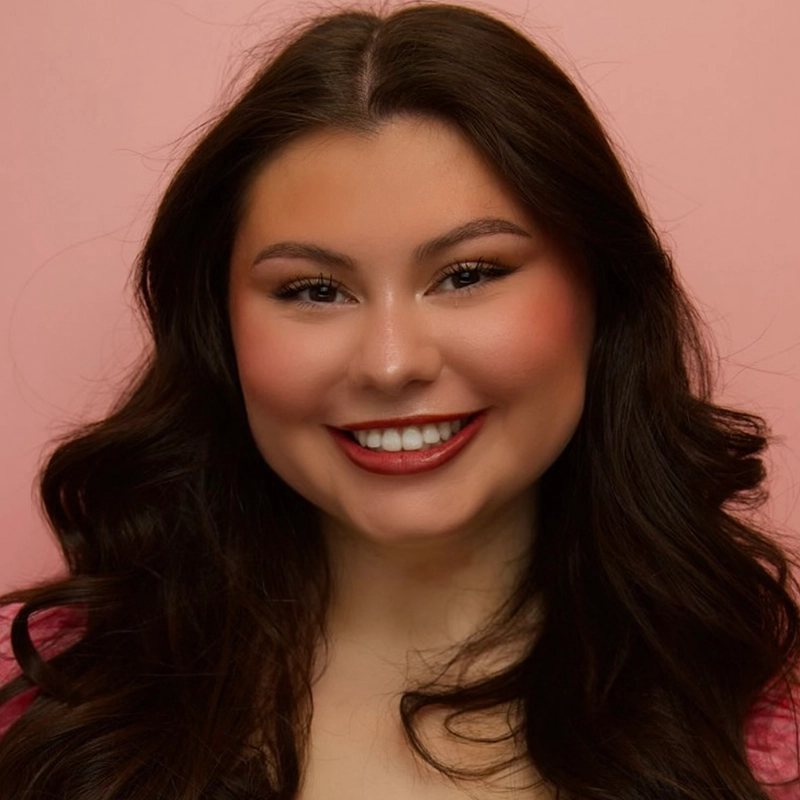 A young woman with long, wavy dark hair smiles at the camera. Mary Grace Smith has a fair complexion, defined eyebrows, winged eyeliner, and red lipstick. She wears a pink top and is posed in front of a soft pink background.