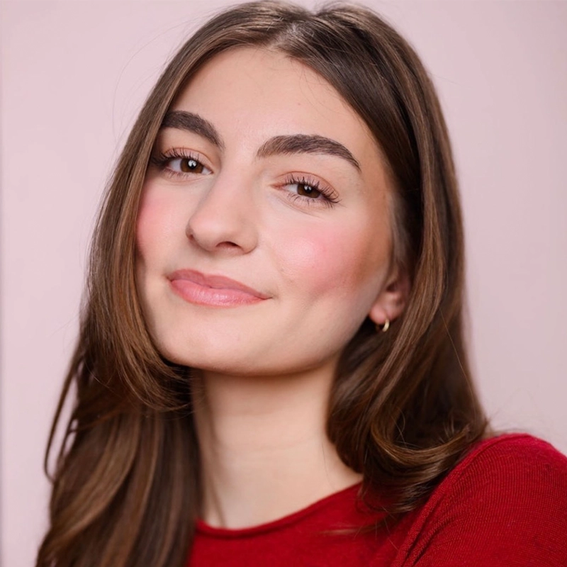 A young woman with long brown hair and full eyebrows smiles softly. She is wearing a red top and small hoop earrings. The background is a soft, light pink, and the lighting is even, highlighting her natural makeup and rosy cheeks.