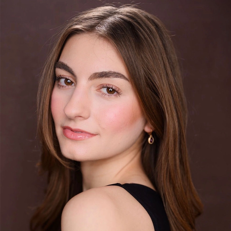 Arielle Ziff, a young woman with long brown hair and arched eyebrows, looks over her shoulder at the camera. She has light skin, natural makeup with rosy cheeks and pink lips, small gold earrings, and a black top against a plain brown background.