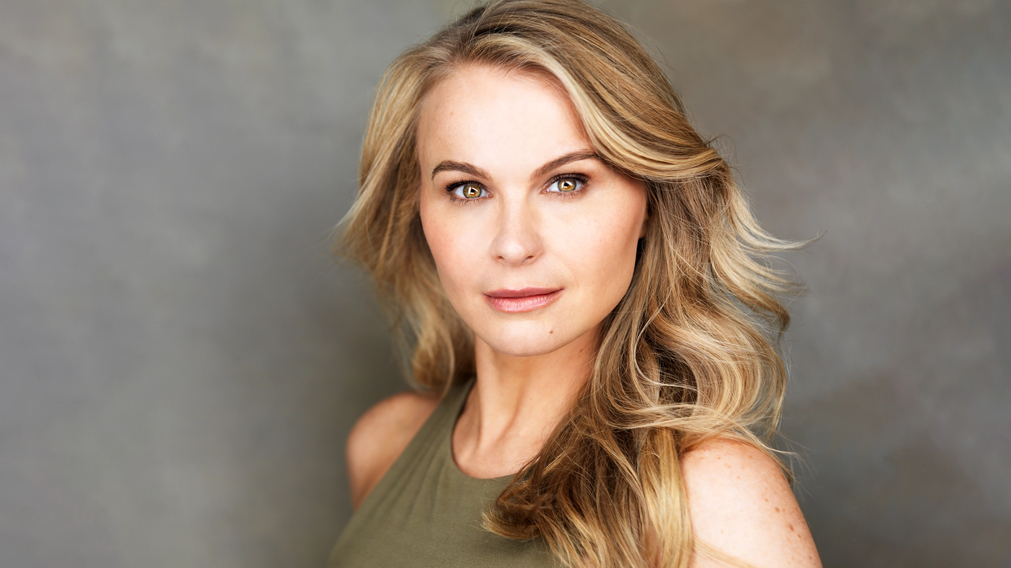 A woman with long, wavy blonde hair and fair skin looks directly at the camera. She has brown eyes, subtle makeup, and wears an olive green sleeveless top. Stacie Bono, known from The Great Escape, is set against a soft gray and brown gradient background.