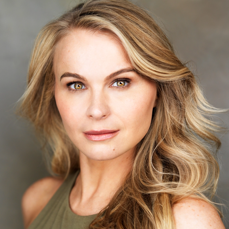 A woman with long, wavy blonde hair, fair skin, and hazel eyes looks directly at the camera. She wears a sleeveless olive-green top. The background is softly blurred in neutral tones, and the lighting highlights her facial features.