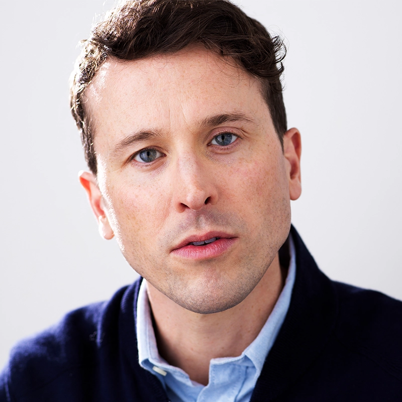 Isaac Woofter, a man with short brown hair and blue eyes, looks directly at the camera with a neutral expression. He wears a light blue collared shirt under a dark sweater against a plain, softly lit background that highlights his face.