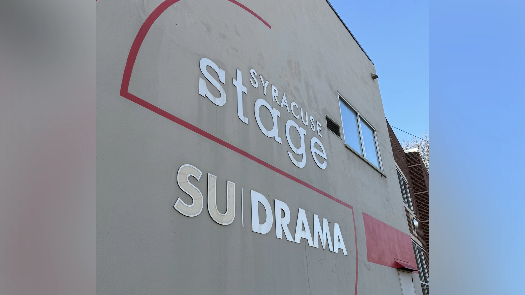 A beige building wall displays large white and metallic letters reading “Syracuse Stage,” “SU Drama,” and highlights Syracuse University's BFA Musical Theatre. Red curved designs accent the text. A clear blue sky and fire escape appear in the background.