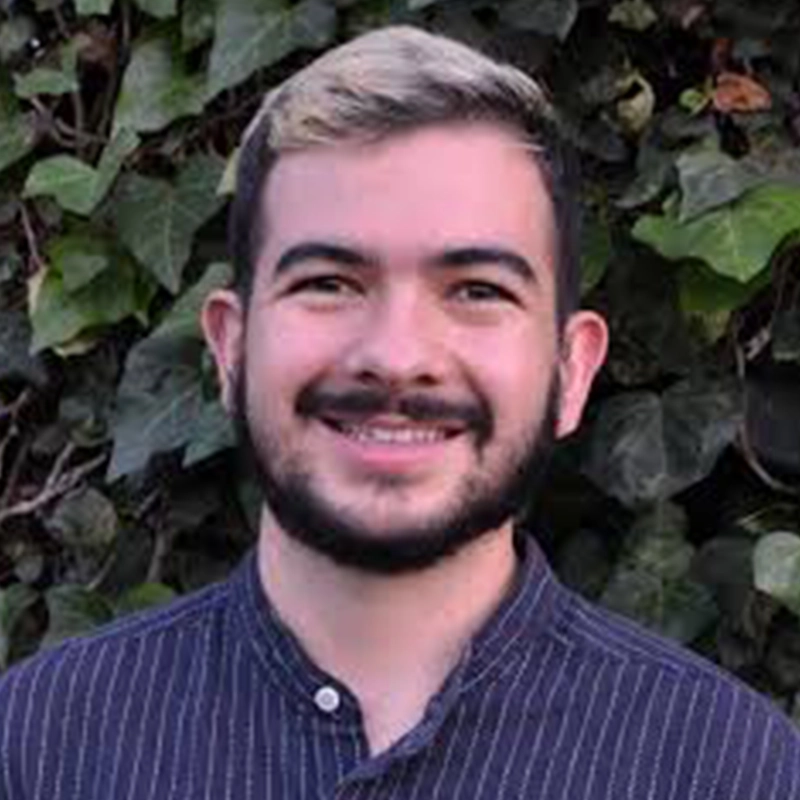 A young man, Timothy Fenton, with short, dark hair and a light streak on top is smiling at the camera. He has a trimmed beard and mustache, and is wearing a dark, striped shirt. Behind him is a leafy, green background.