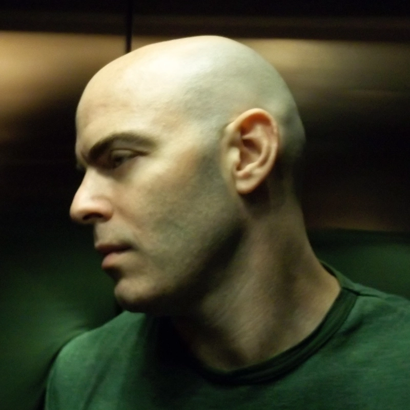 A bald man in a green shirt, possibly Timothy Fenton, is shown in profile, looking down and to the left. The background is dark and blurred, creating a moody atmosphere. Soft lighting highlights the contours of his head and face.