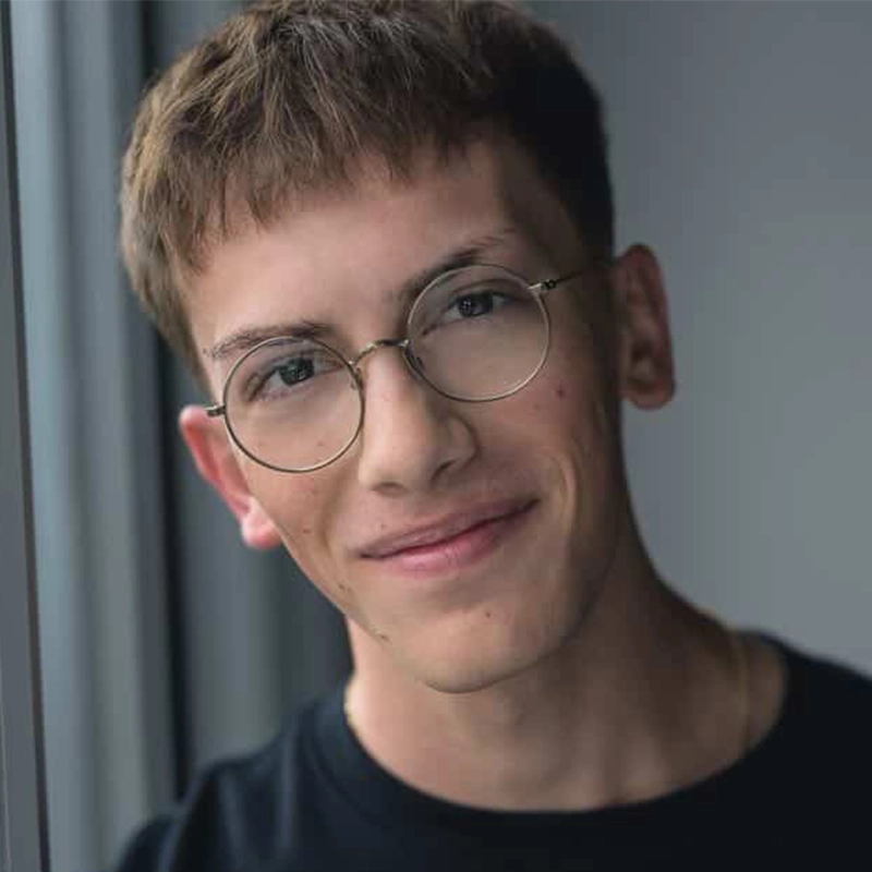 A young man with short brown hair and round glasses smiles gently while looking into the camera. Isaac Woofter wears a black shirt, and natural light from a nearby window softly illuminates his face against a blurred, neutral background.