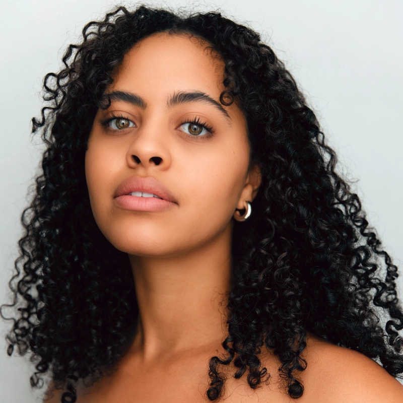 Morgan Wardlaw, a woman with medium brown skin and curly dark hair, looks at the camera. She has light green eyes, full lips, and wears small hoop earrings. The background is plain and light, and her expression is calm and neutral.
