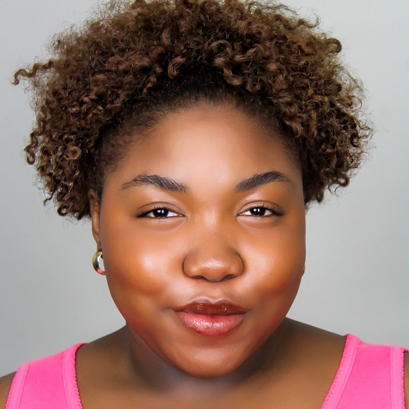 Jyonnah Ware, with medium-dark skin and curly brown hair, smiles softly. Wearing a pink top and small hoop earrings, she stands against a plain light gray background that highlights her calm, confident expression.