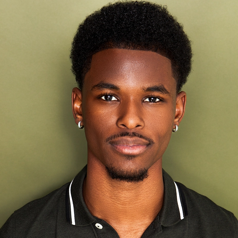 A young Black man with short, curly hair and trimmed facial hair faces the camera against an olive green background. He wears a black collared shirt with white trim and small hoop earrings, and has a neutral, confident expression.