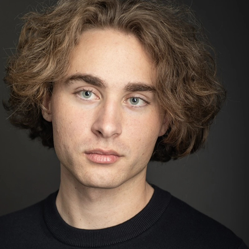 A young man with fair skin, wavy light brown hair, and blue eyes stares softly at the camera. He wears a dark crew-neck sweater and is set against a plain, dark background. The lighting highlights his facial features.
