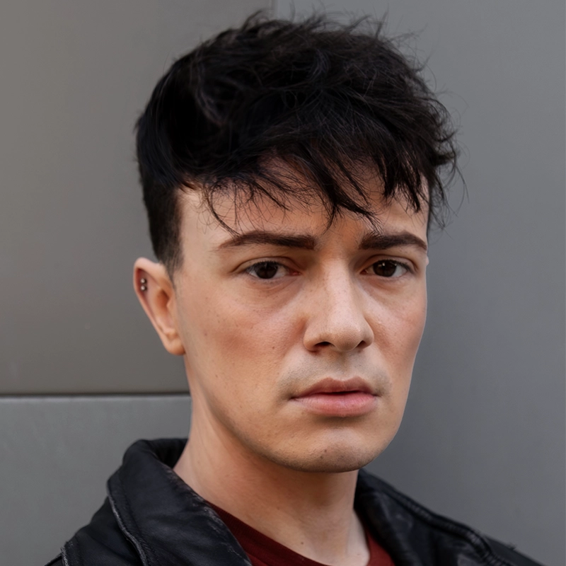 A young person with short, tousled black hair and fair skin looks directly at the camera with a neutral expression. They wear a black leather jacket over a dark red shirt and have an ear piercing. The background is a plain gray wall.