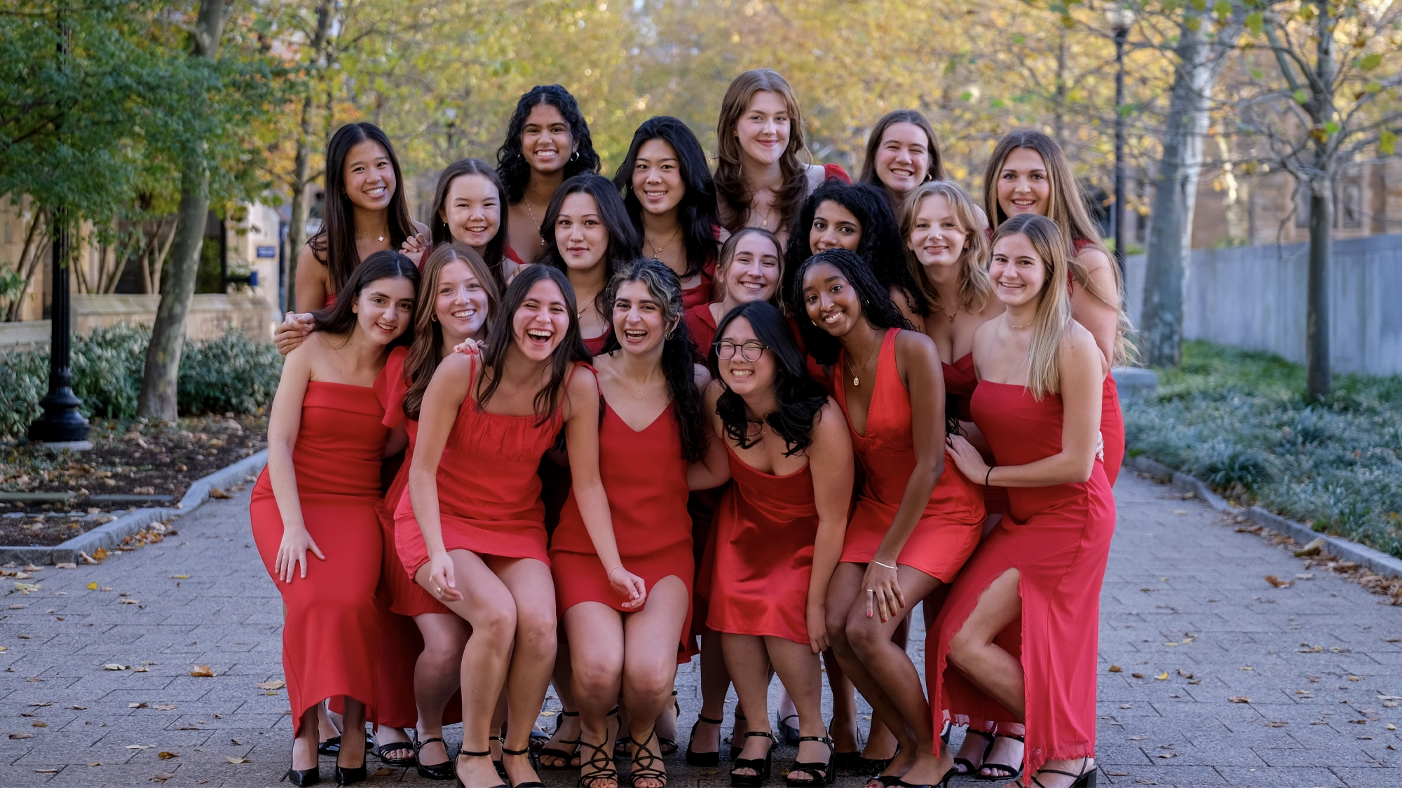 A group of nineteen young women stand closely together on a stone walkway, smiling and posing for a photo in vibrant red dresses at Yale University. Behind them, autumn trees with yellow leaves add warmth to the scene.