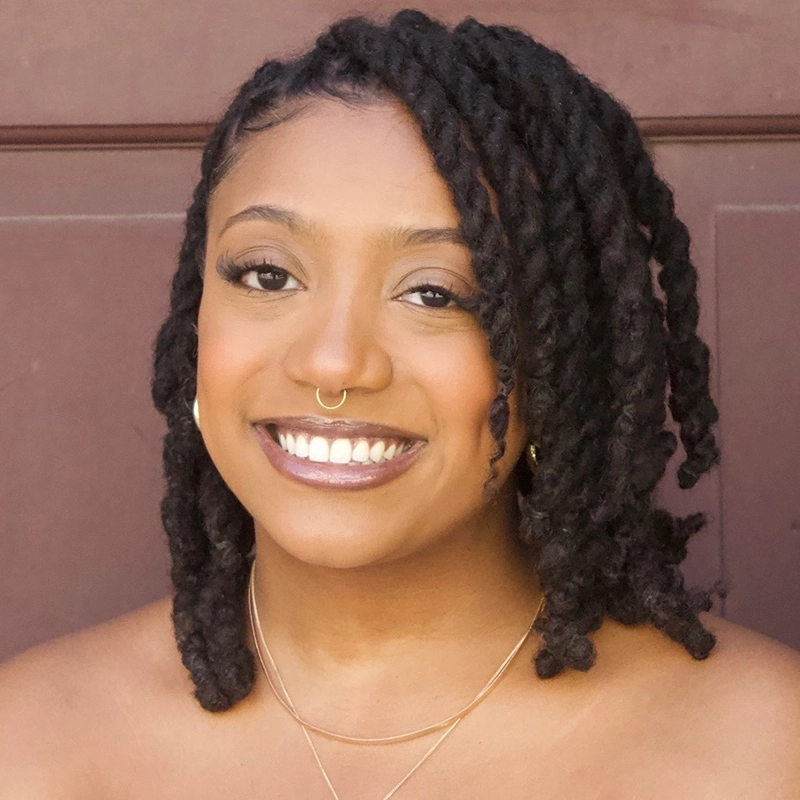 Zeleah Emmanuel smiles with dark, twisted shoulder-length hair and a gold septum ring, posing in front of a brown background. She wears natural makeup, nude lipstick, layered gold necklaces, and her shoulders are bare.