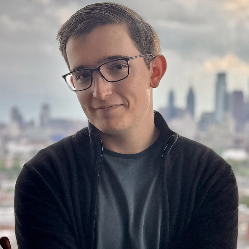 A young person with short brown hair, wearing glasses, a dark zip-up sweater, and a gray t-shirt, stands in front of a window with a blurred city skyline under a cloudy sky. Val Zvinyatskovsky smiles slightly, arms loosely crossed.