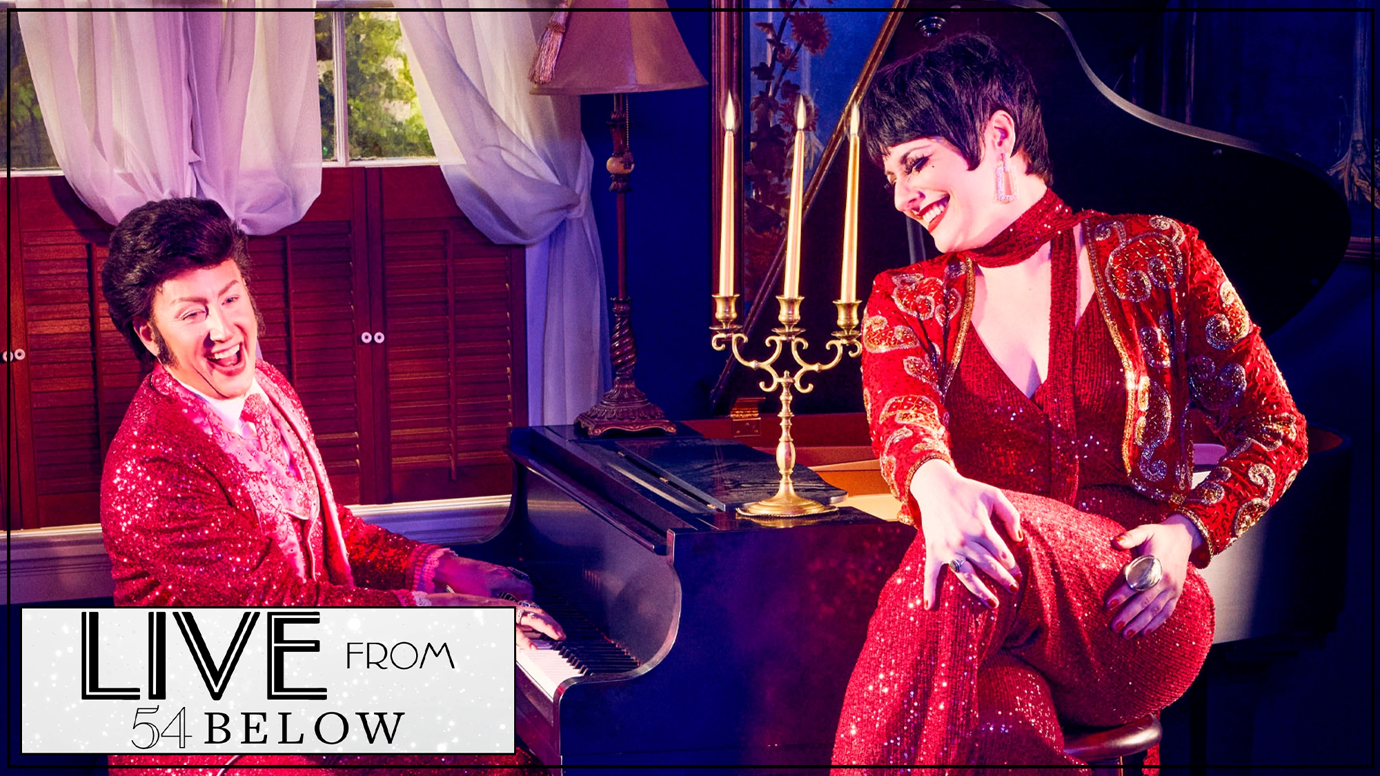 Two performers in sparkling red outfits sit at a black grand piano in a glamorous Liberace tribute. One plays, smiling broadly; the other poses atop the piano. A candelabra, lamp, and sheer curtains adorn the warm, elegant room. 