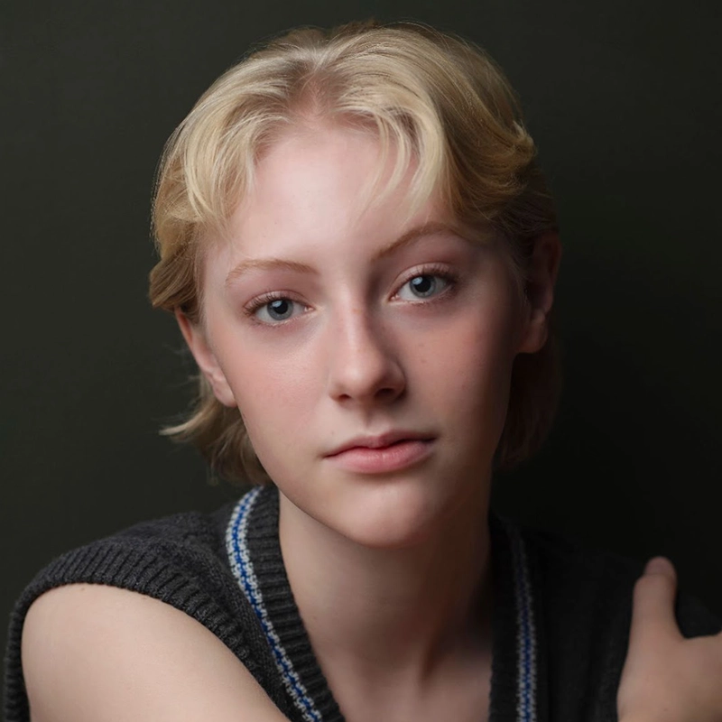 A person with short blond hair and light skin gazes gently at the camera. They wear a sleeveless dark sweater with a blue and white stripe. The background is a muted dark green, and the lighting softly highlights their facial features, creating an intimate portrait reminiscent of Asher Alcantara’s understated style.