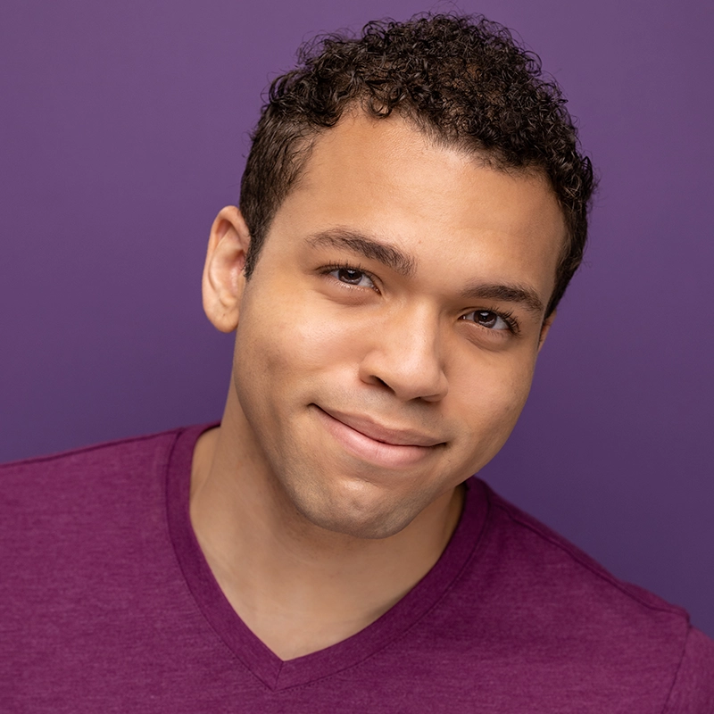 Aaron Valentine, a young man with short curly hair, smiles softly in a purple V-neck shirt. He is posed in front of a matching purple background, giving the photo a harmonious, monochromatic look.