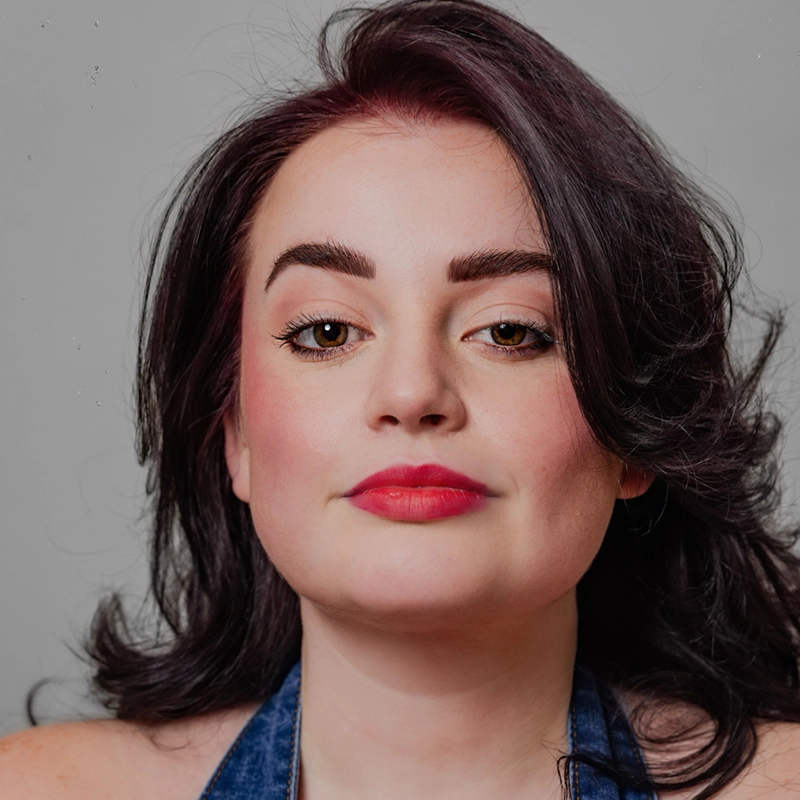 A woman with fair skin, wavy dark brown hair, and bold eyebrows looks confidently at the camera. She wears pink lipstick, subtle eye makeup, and a blue halter top. The background is plain and gray.