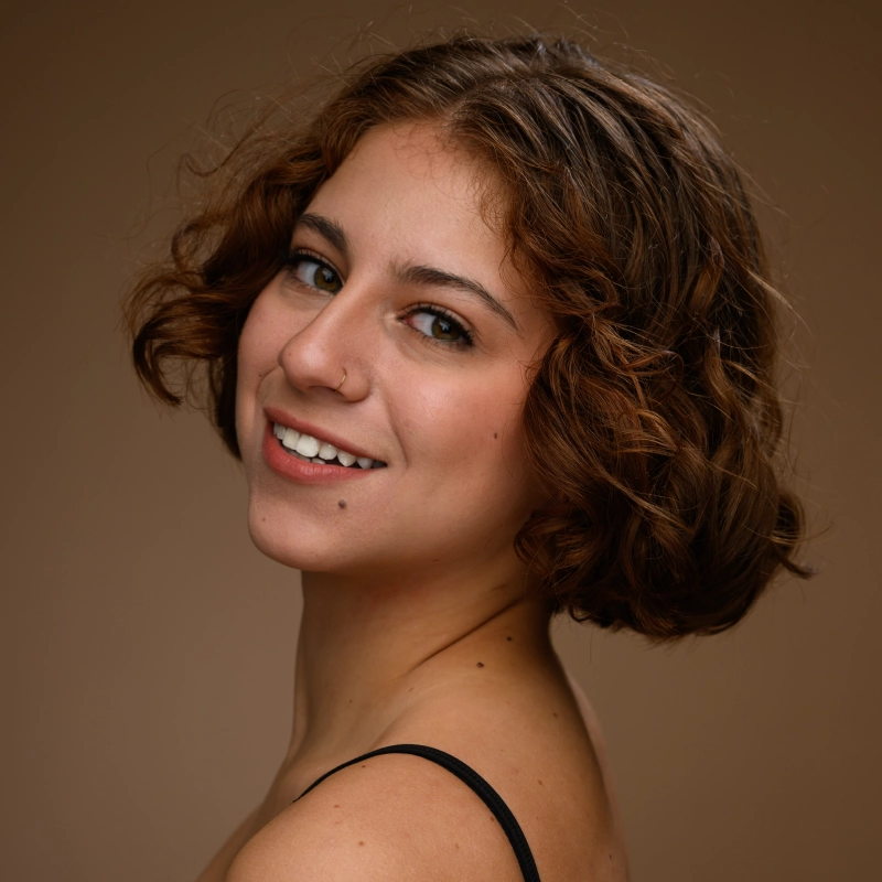 A young woman with short, curly brown hair smiles at the camera. She has a nose ring and a small mole on her upper lip. She wears a black strap top and is posed against a plain brown background. The lighting is soft and warm.