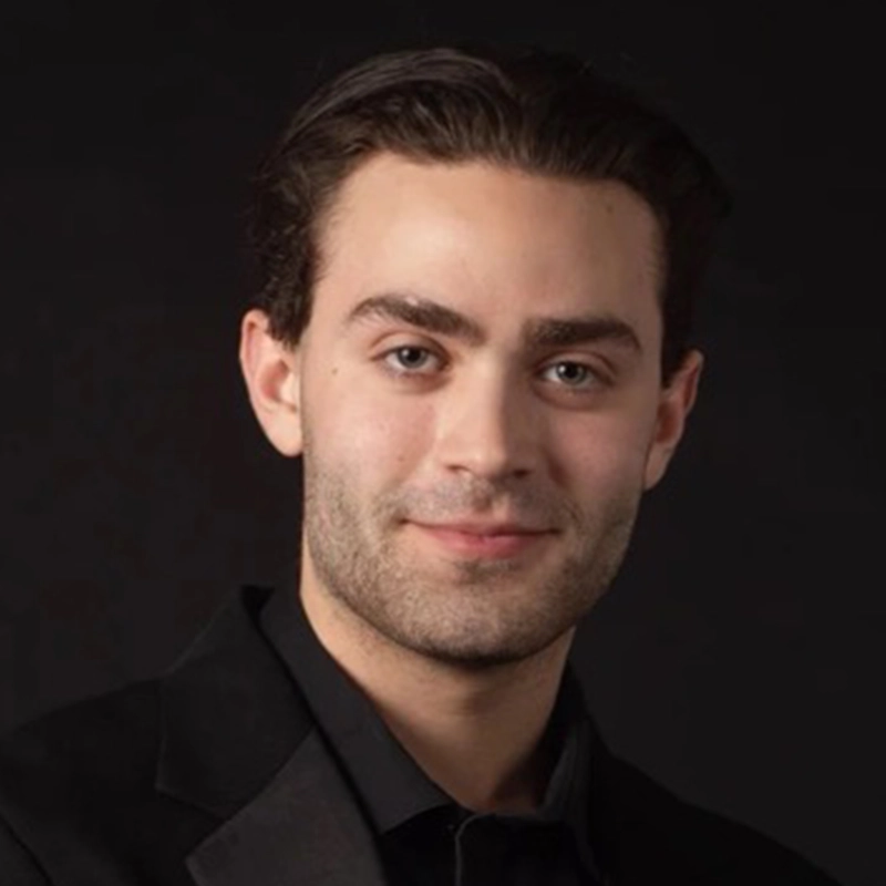 A young man with fair skin, dark hair, and subtle stubble smiles slightly. Gaël Mouton-Sady wears a black suit jacket over a black shirt. The background is solid black, creating a formal and professional portrait atmosphere.