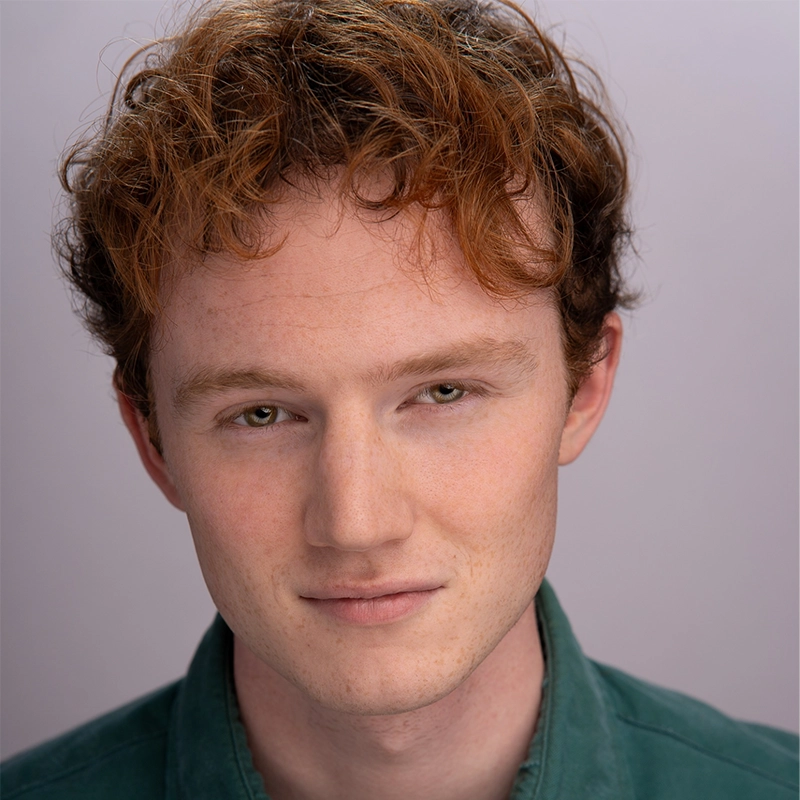 A young person with fair skin, short curly red hair, and greenish eyes gazes at the camera with a calm, slight smile. They are wearing a forest green collared shirt from Surfac. The background is plain and softly lit in light lavender-gray tones.