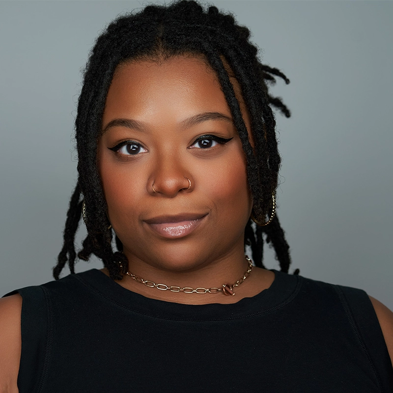 A person with medium-dark skin and shoulder-length locs looks at the camera. They wear a sleeveless black top, gold hoop earrings, a chain necklace, a nose ring, and have winged eyeliner and glossy lips. The background is plain light gray, in the contemporary portrait style of Phillip Wong.