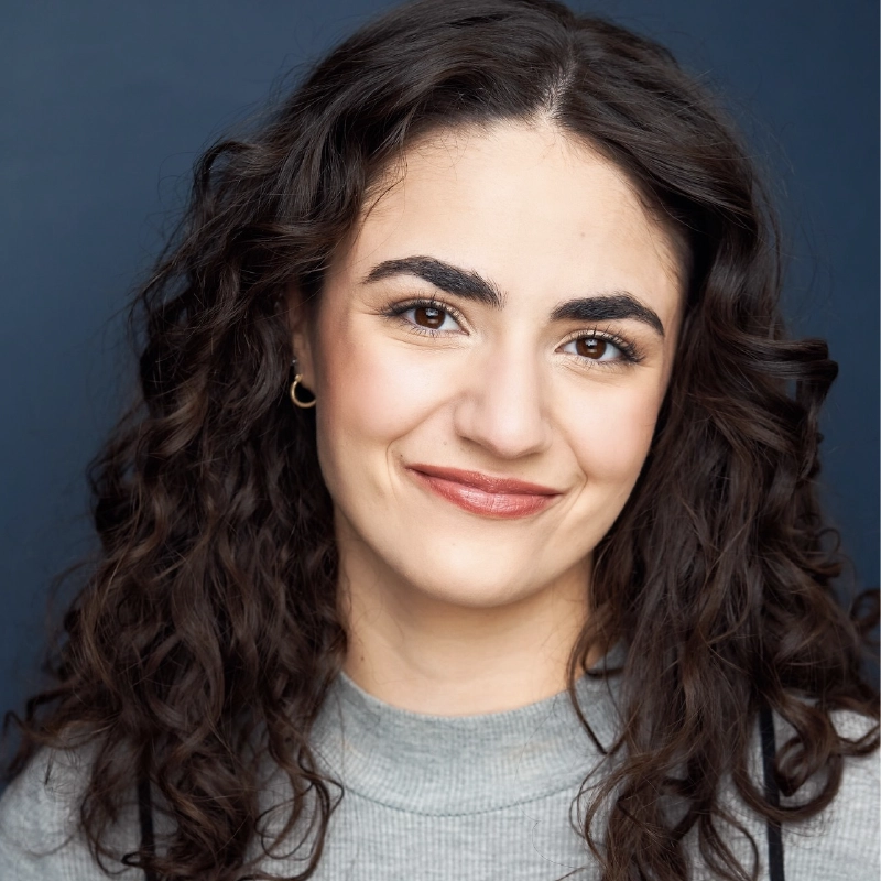 A woman with long, dark curly hair and thick eyebrows smiles slightly. She wears a light gray top, gold hoop earrings, and natural makeup. The background is a solid dark blue. The image is well-lit, focusing on the face and shoulders of Alexandra Zografos.