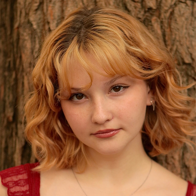 A young person with wavy, chin-length, light brown hair and bangs stands in front of a tree trunk. They wear a red lace top, small earrings, and a subtle necklace. Their expression is neutral, with soft makeup and a gentle look into the camera, capturing the understated elegance reminiscent of Jenny Walker’s signature style.