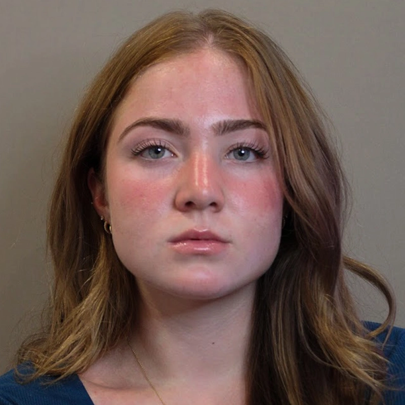 A young woman with light skin, long brown hair, and blue eyes gazes directly at the camera with a neutral expression. Martina Balduzzi is wearing a blue top and small hoop earrings; her cheeks are slightly flushed. The background is plain and beige.