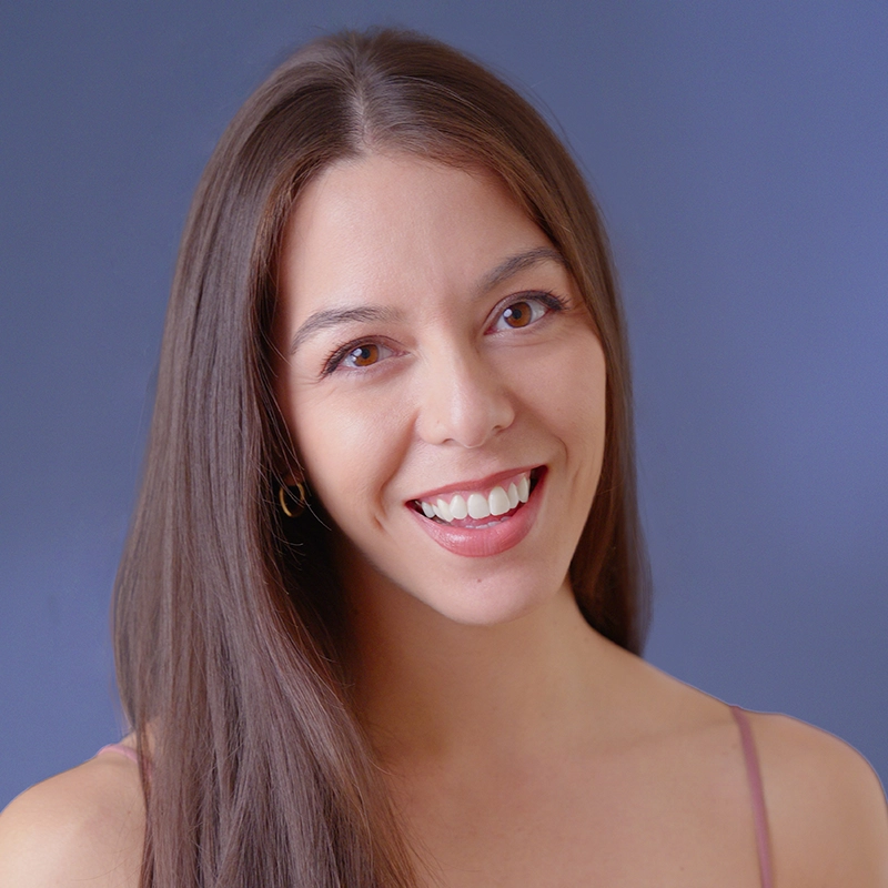 A young woman with long straight brown hair smiles warmly at the camera. She has light skin, brown eyes, and is wearing a sleeveless, thin-strap top. The background is a solid blue color.