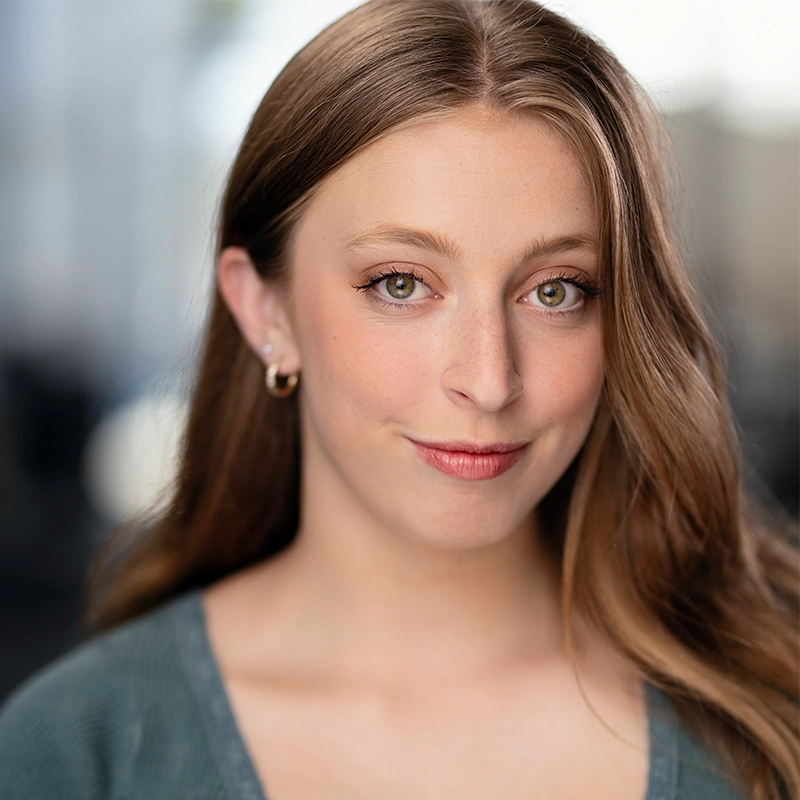 Casey Keeler, a young woman with long light brown hair, hazel eyes, and fair skin, looks at the camera, smiling softly. She wears small hoop earrings and a sage green top. The blurred background is bright and neutral.