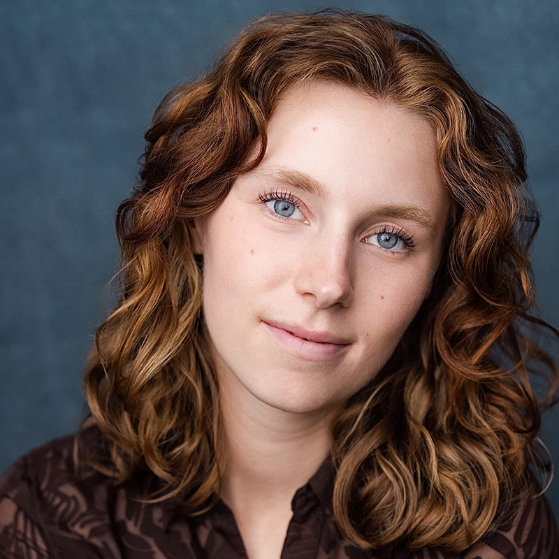 A young person with wavy, shoulder-length brown hair and blue eyes smiles softly. They wear a dark, patterned top and are posed in front of a textured blue background. The lighting is soft, highlighting their clear skin and natural expression.
