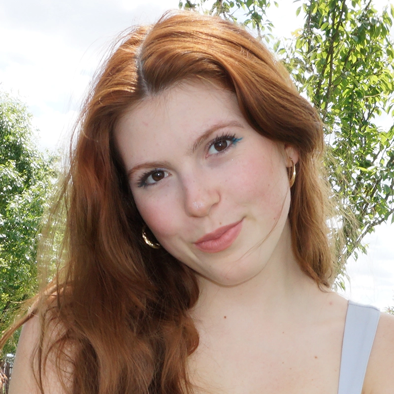 A young woman with long auburn hair and fair skin smiles slightly. She wears hoop earrings and a light blue top. Green trees and a partly cloudy sky are in the background, suggesting an outdoor setting—perhaps reminiscent of the serene moments often captured by Gaël Mouton-Sady.