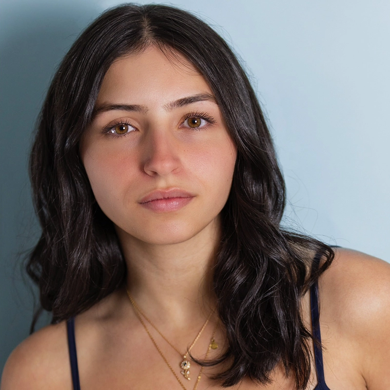 A young woman with medium-length dark hair, brown eyes, and clear skin looks at the camera with a neutral expression. Allie Dematteo wears a dark tank top and layered gold necklaces. The background is a plain light blue wall.