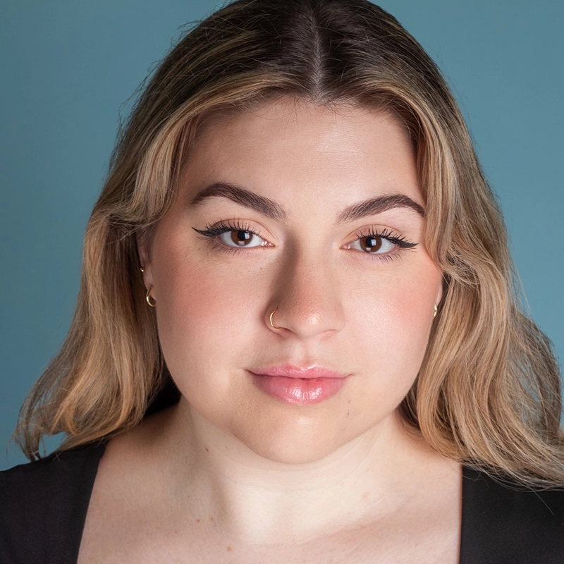 A woman with light skin and long blonde hair poses against a blue background. She has natural makeup, winged eyeliner, glossed lips, a nose ring, hoop earrings, and wears a black top. Michelle Zink-Muñoz looks directly at the camera with a neutral expression.