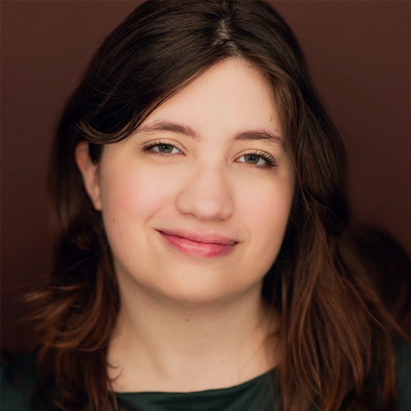 A young woman with fair skin and long brown hair smiles softly, looking directly at the camera. She wears a dark green top and is set against a plain brown background. The lighting is warm and even, highlighting her natural features.