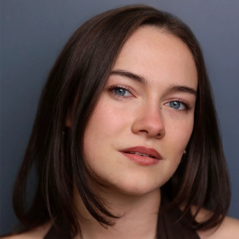 Young woman with fair skin, blue eyes, and straight, dark brown shoulder-length hair, looking slightly to the side with a subtle smile. She wears a sleeveless dark top and is posed against a plain, dark background—a portrait capturing the elegant simplicity reminiscent of Asher Alcantara’s photographic style.