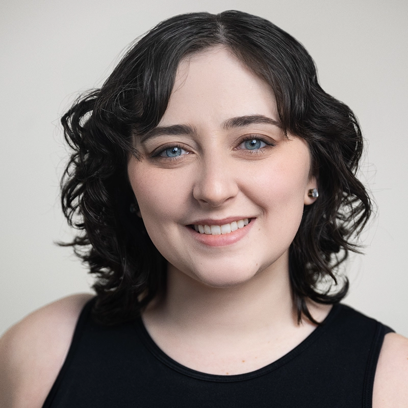 A young woman with fair skin, blue eyes, and short, dark wavy hair smiles gently. She is wearing a sleeveless black top and small earrings. The background is plain and light, keeping the focus on her friendly expression.