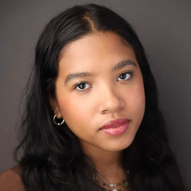 A young woman with long black wavy hair, wearing silver hoop earrings and a beaded necklace, gazes seriously at the camera. She has warm brown skin, soft pink lips, and natural makeup. The background is plain and gray, allowing her expression to quietly say: Call Me Katie.