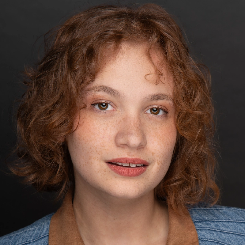 Jayline Ross, a young person with light skin, freckles, and short curly auburn hair, looks at the camera with a soft smile. They wear a blue top with a brown collar. The dark background makes Jayline’s features stand out.
