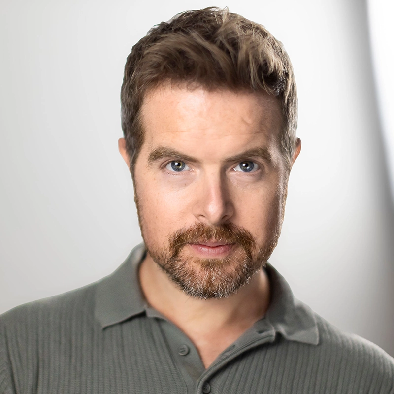 A man with light skin, short brown hair, and a trimmed beard looks directly at the camera. He is wearing a green collared shirt, and the background is softly blurred in shades of white and gray, creating a professional headshot appearance.
