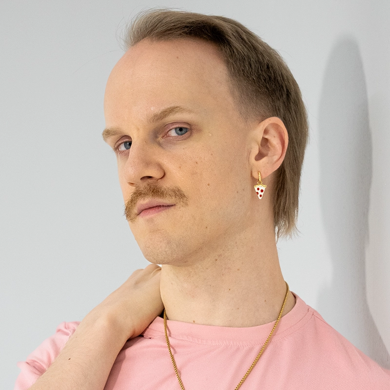 Aaron Valentine, with short light brown hair and a mustache, looks at the camera. Wearing a light pink shirt, gold necklace, and a pizza-shaped earring, his hand rests on his neck against a plain light grey background.