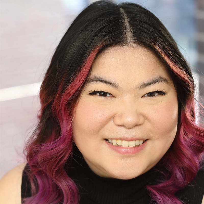Michelle Zink-Muñoz, a woman with long, black hair featuring pink tips, smiles at the camera. She has fair skin, black eyebrows, and wears a black sleeveless turtleneck. The background is softly blurred with bright natural light.
