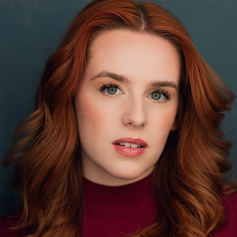 A woman with long wavy red hair and fair skin looks at the camera. She has light makeup, subtle blush, and natural lips. She wears a burgundy top and is posed in front of a dark, plain background. The lighting is soft and highlights her facial features.