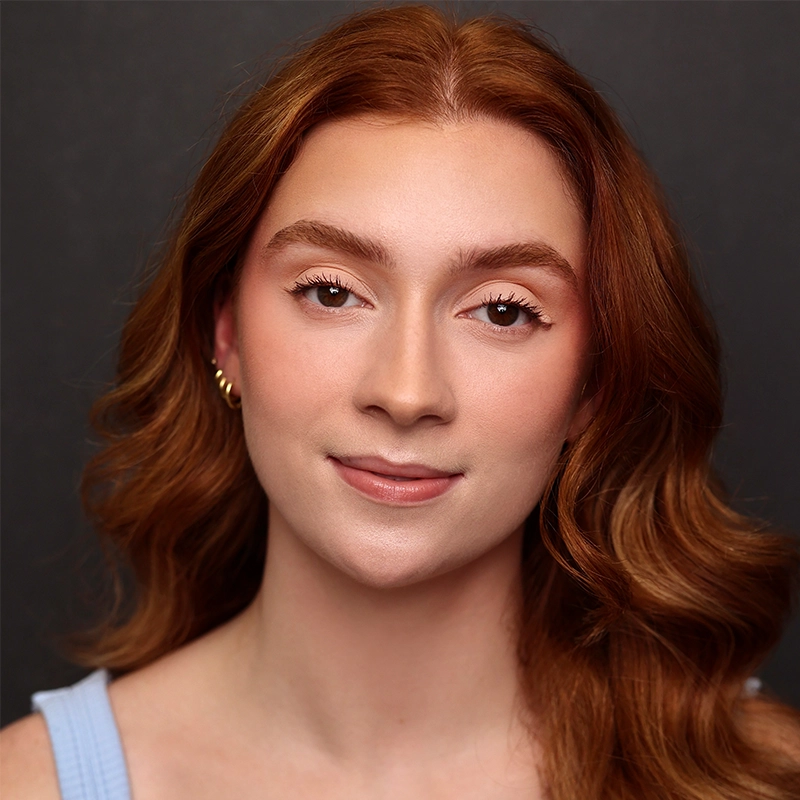A young woman with long, wavy auburn hair poses against a dark background. She has fair skin, light makeup, and wears a light blue top and small gold hoop earrings. Megan Ludwig looks directly at the camera with a calm, gentle smile.