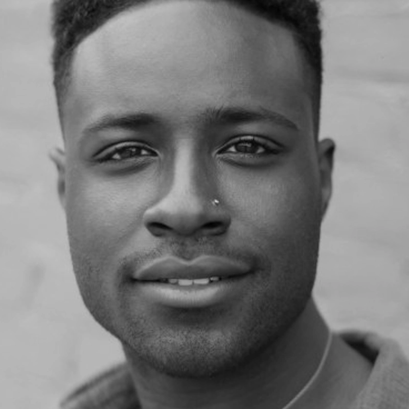 A young man with short, curly hair and a neatly groomed beard looks at the camera. He has a small nose piercing and wears a collared shirt. The background is blurred, and the photo is in black and white, capturing Murphy Lorenzo Applin Jr.’s poised expression in timeless style.