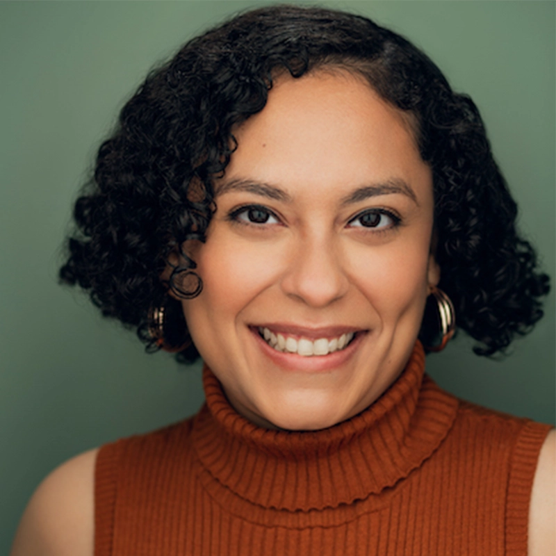 Smiling person with medium brown, curly hair wears hoop earrings and an orange sleeveless turtleneck. The background is a muted green, and soft lighting highlights their friendly expression.