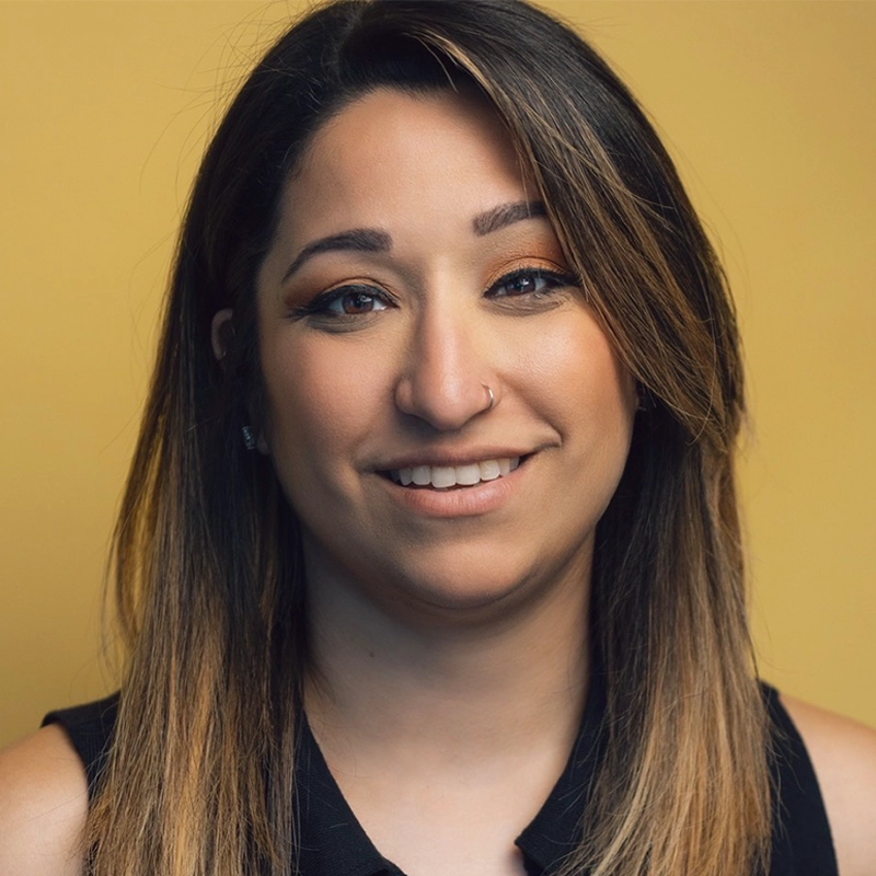 A woman with straight, shoulder-length brown hair and a nose ring smiles gently at the camera. She wears a sleeveless black top and has natural makeup. The background is a plain, warm yellow, perfectly complementing the inviting vibe of this 