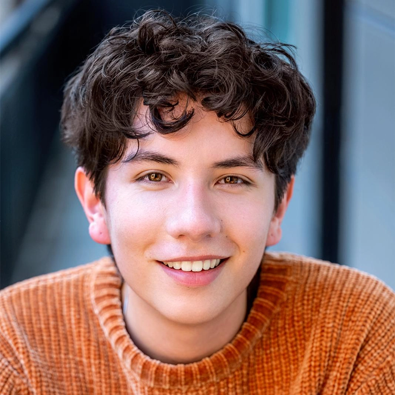 A young person with short, curly brown hair smiles at the camera. They wear a ribbed, orange sweater. The background is blurred, showing hints of a building and railing, with soft natural light on their face—capturing Asher Alcantara’s warm and approachable presence.