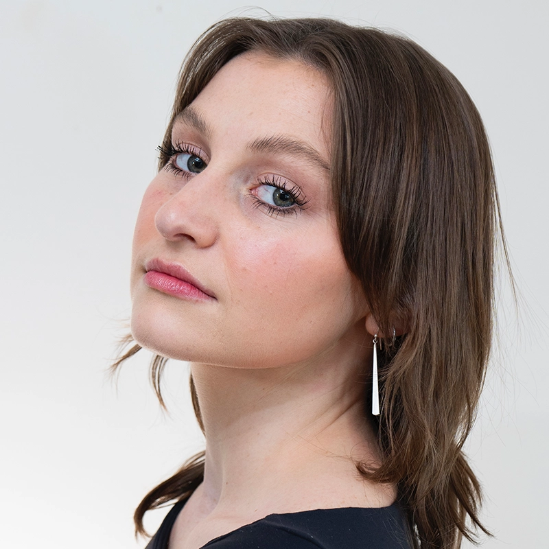 Gabrielle Farley, a woman with light skin and straight brown hair, looks over her shoulder at the camera. She wears a black top, subtle makeup, long silver earrings, and has a neutral expression. The background is plain white.